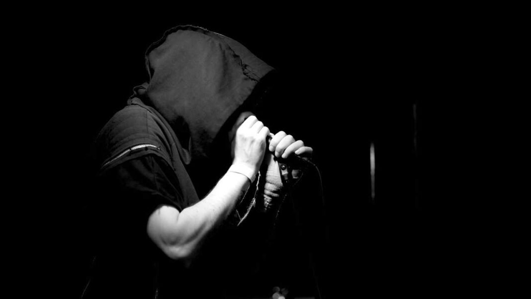 Persona con capucha sostiene un micrófono junto a la boca en una imagen en blanco y negro sobre fondo oscuro.