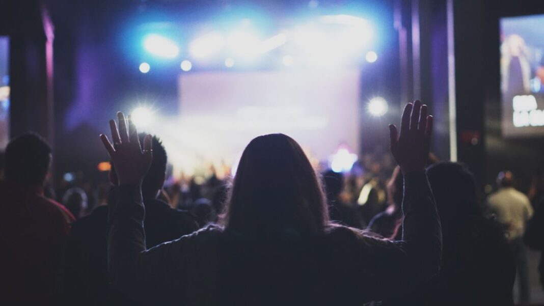 Público de espaldas en un concierto, con una persona en primer plano que levanta ambos brazos frente a un escenario iluminado.