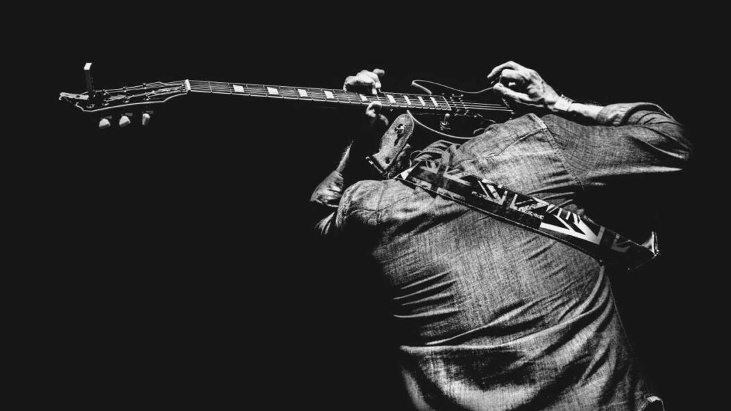 Fotografía en blanco y negro de una persona de espaldas tocando un bajo eléctrico, con camisa de mezclilla y correa estampada, sobre fondo negro.