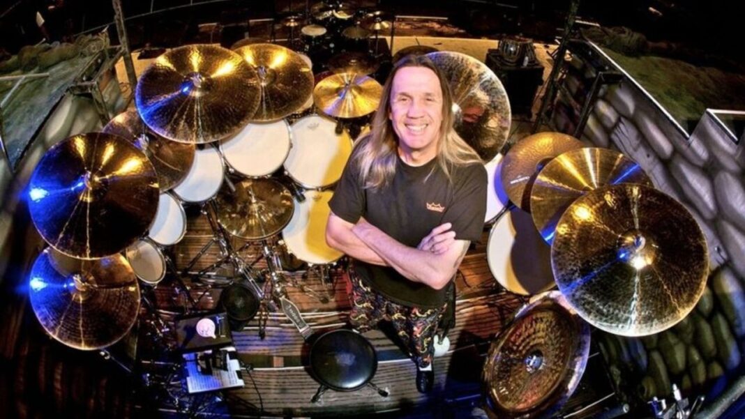 Baterista de cabello largo y camiseta negra sonríe con los brazos cruzados rodeado de una gran batería con platillos dorados en un escenario.