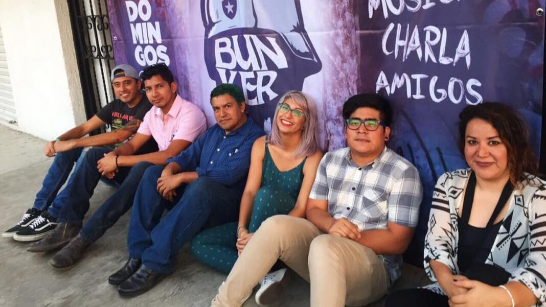 Seis personas sentadas en el suelo junto a una pared con un cartel morado que muestra texto y un dibujo de una cabeza.