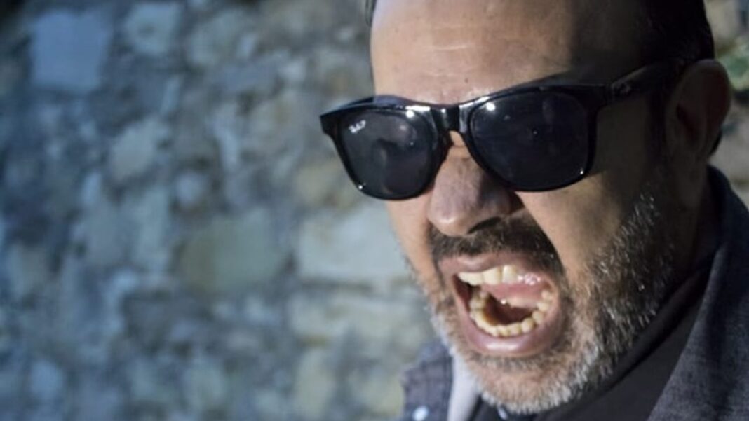 Primer plano de un hombre calvo con barba y gafas de sol negras, con la boca abierta mostrando los dientes, frente a una pared de piedra desenfocada.