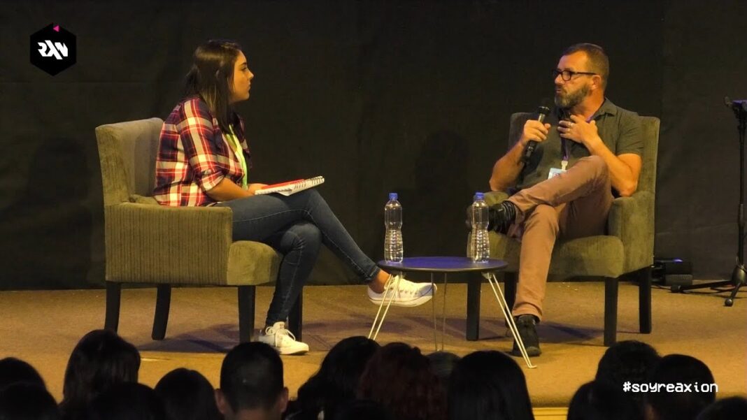 Dos personas sentadas en sillones sobre un escenario; una mujer con camisa de cuadros y un hombre con micrófono, ante un público.