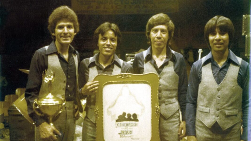 Cuatro hombres de pie en interiores, con camisas oscuras y chalecos claros, sostienen un trofeo dorado y una placa enmarcada.