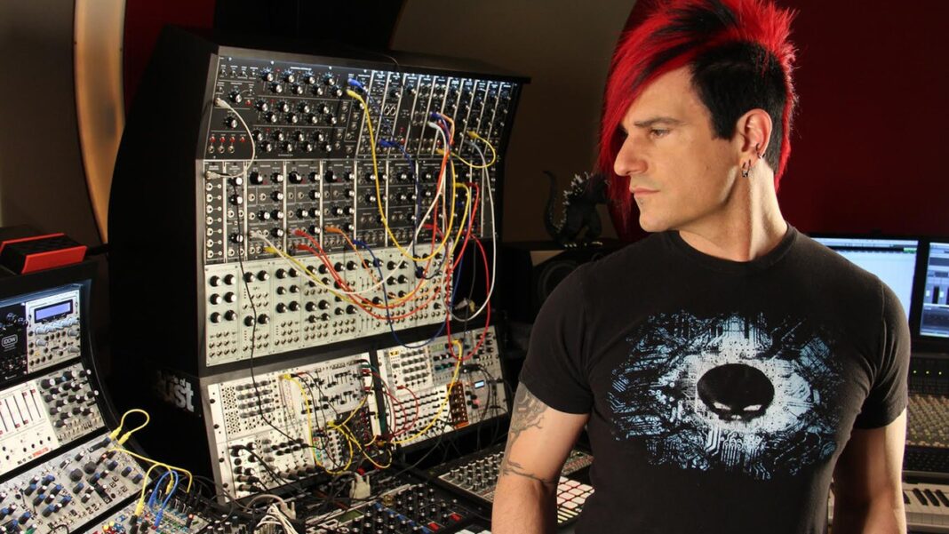 Un hombre de cabello negro con mechones rojos y camiseta negra estampada observa de lado junto a un gran panel modular lleno de cables y controles.