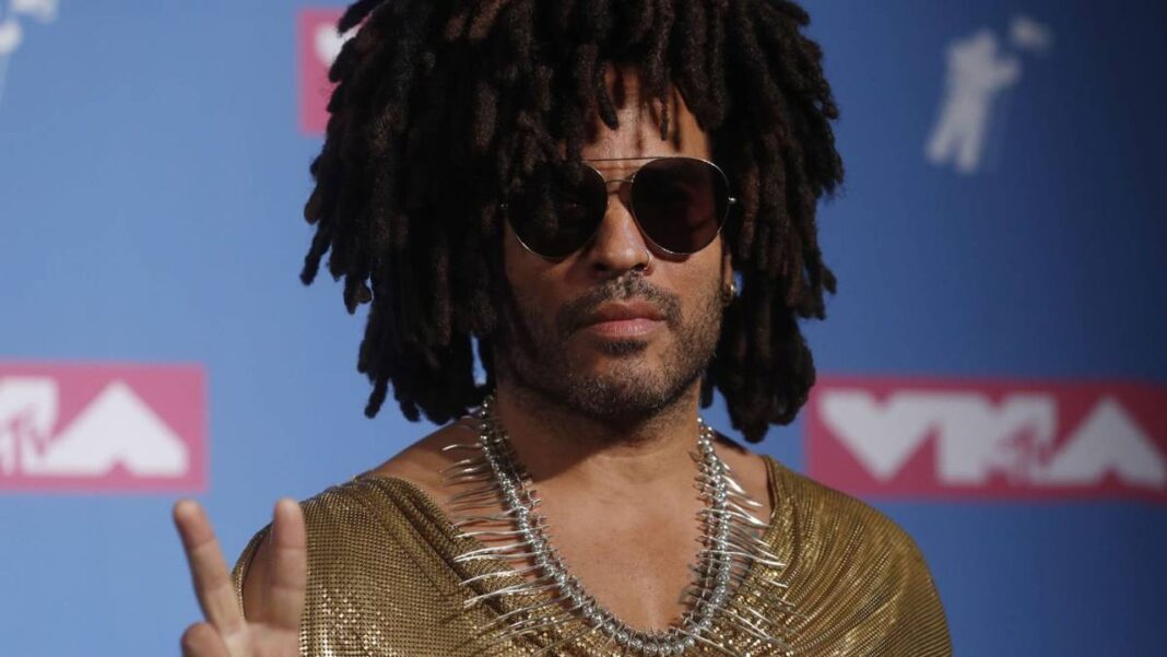 Hombre con rastas largas, gafas de sol y collar de pinchos hace el signo de paz frente a un fondo azul con logotipos.