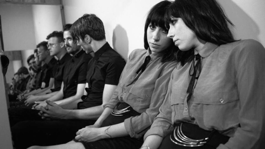 Fotografía en blanco y negro de personas sentadas en fila contra una pared; dos mujeres con flequillo y blusas claras en primer plano y varios hombres de camisa oscura detrás.