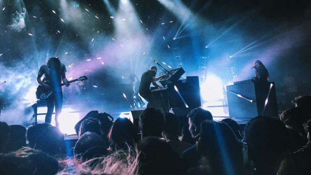 Concierto en vivo con tres músicos en el escenario, luces azules, humo y chispas, y el público de espaldas en primer plano.