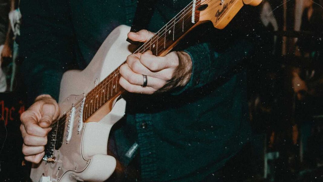 Primer plano de las manos de una persona con anillo tocando una guitarra eléctrica blanca de cuerpo claro, sobre una camisa oscura y fondo desenfocado.