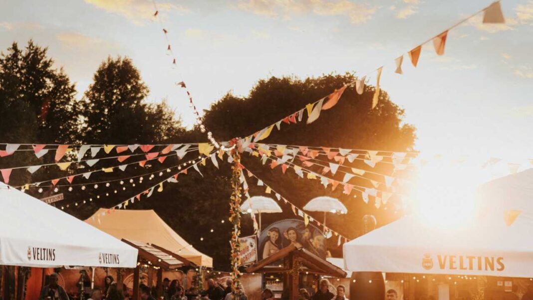 Carpas blancas con el rótulo Veltins, banderines de colores y luces colgantes sobre una multitud en un recinto al aire libre al atardecer.