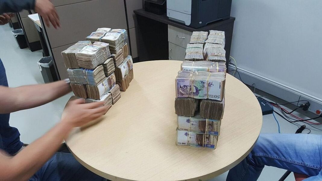 Dos grandes pilas de fajos de billetes se alzan sobre una mesa redonda de madera clara en una oficina, con brazos y manos de personas cerca.