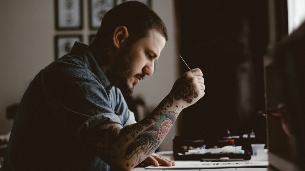 Hombre joven con barba y brazo tatuado, de perfil, sostiene una herramienta fina y trabaja concentrado junto a una mesa.