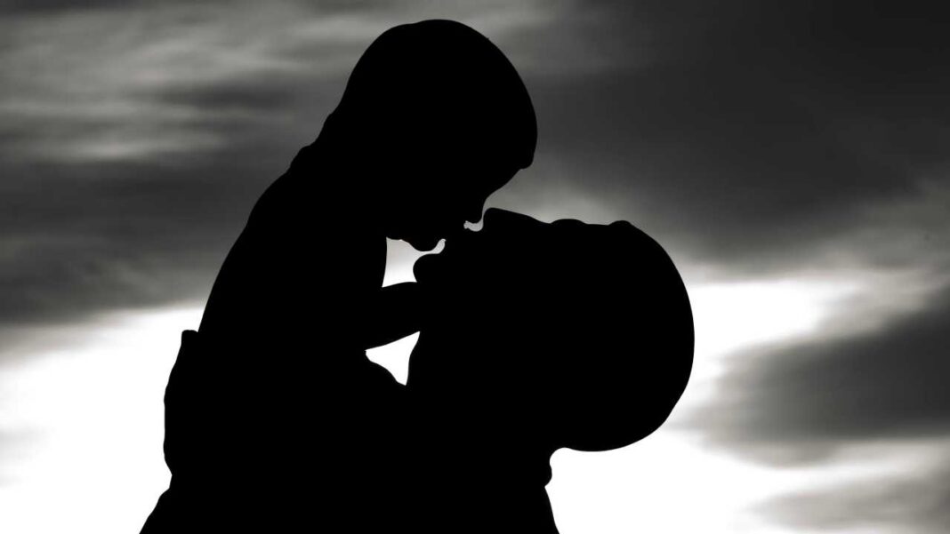Silueta negra de dos personas muy juntas frente a un cielo nublado con luz brillante detrás, una inclinada hacia la otra.