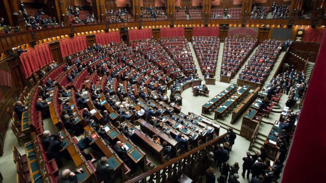 Amplia sala semicircular con filas de asientos rojos, escritorios verdes y numerosas personas sentadas, rodeada por balcones de madera y cortinas rojas.