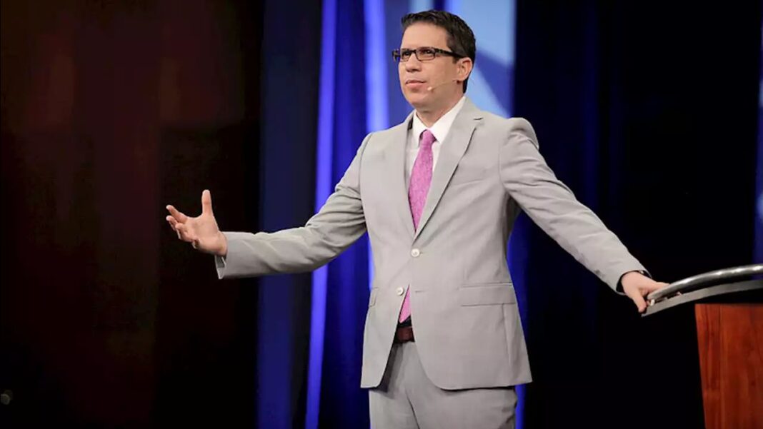 Hombre con traje gris, corbata rosa y gafas habla en un escenario con cortinas azules y un atril de madera.