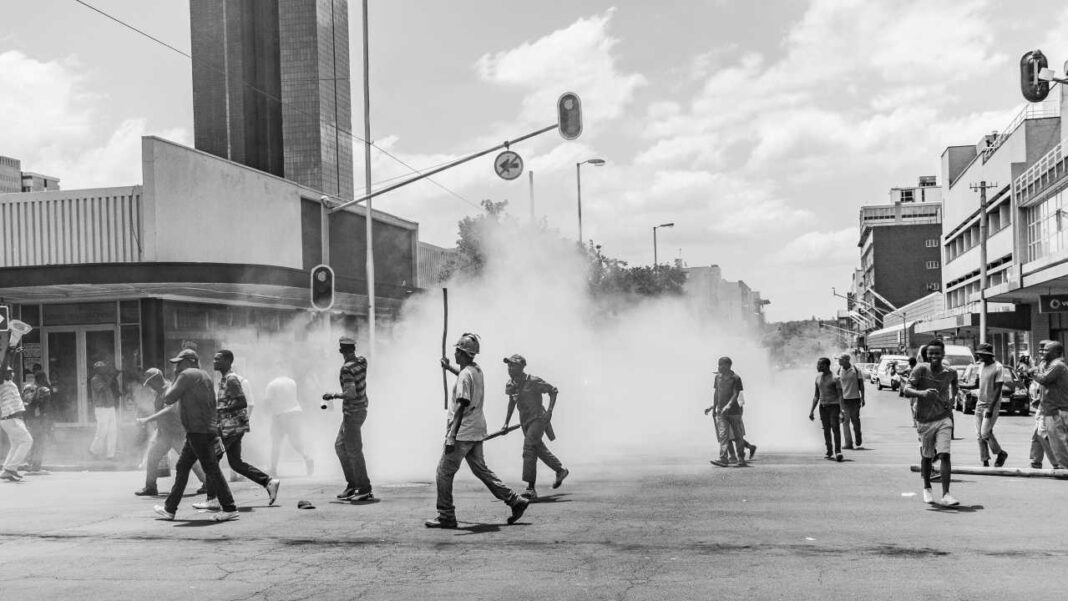 Imagen en blanco y negro de una calle urbana con varias personas caminando y corriendo entre una gran nube de humo, semáforos y edificios al fondo.