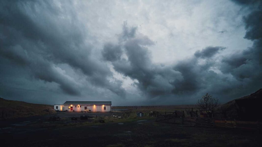 Paisaje rural bajo un cielo tormentoso y nubes oscuras, con una casa blanca iluminada y terreno abierto en penumbra.