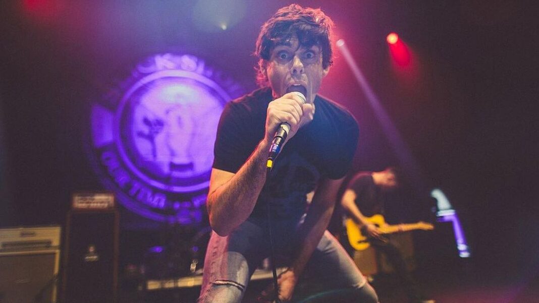 Cantante de pelo oscuro sostiene un micrófono y canta inclinado en un escenario, con luz morada y roja y un músico con guitarra al fondo.