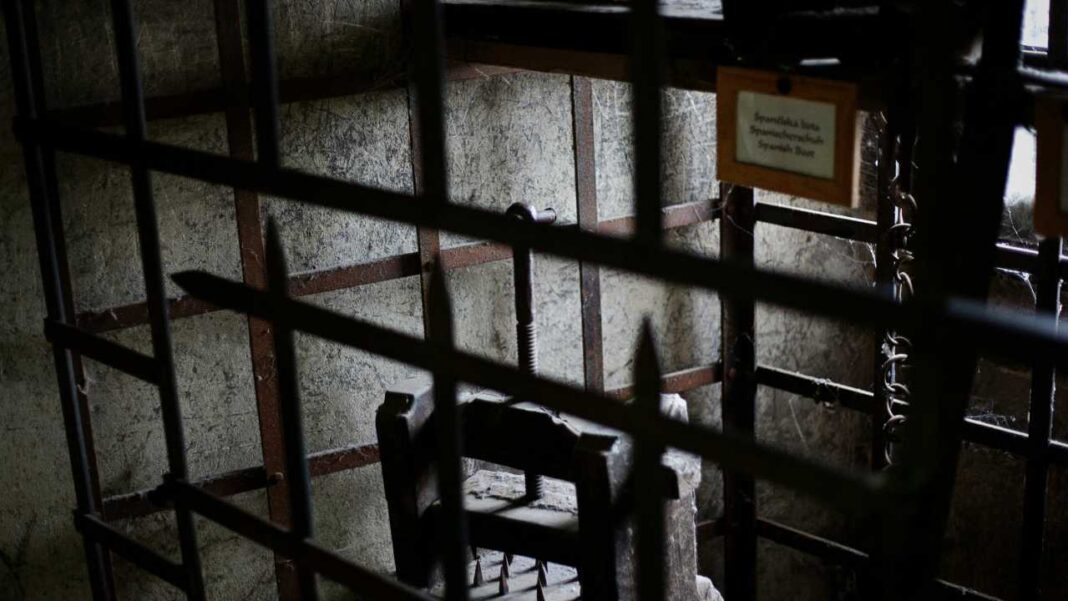 Vista a través de gruesos barrotes metálicos de una estancia oscura con una silla antigua de madera y un cartel colgado en la pared.