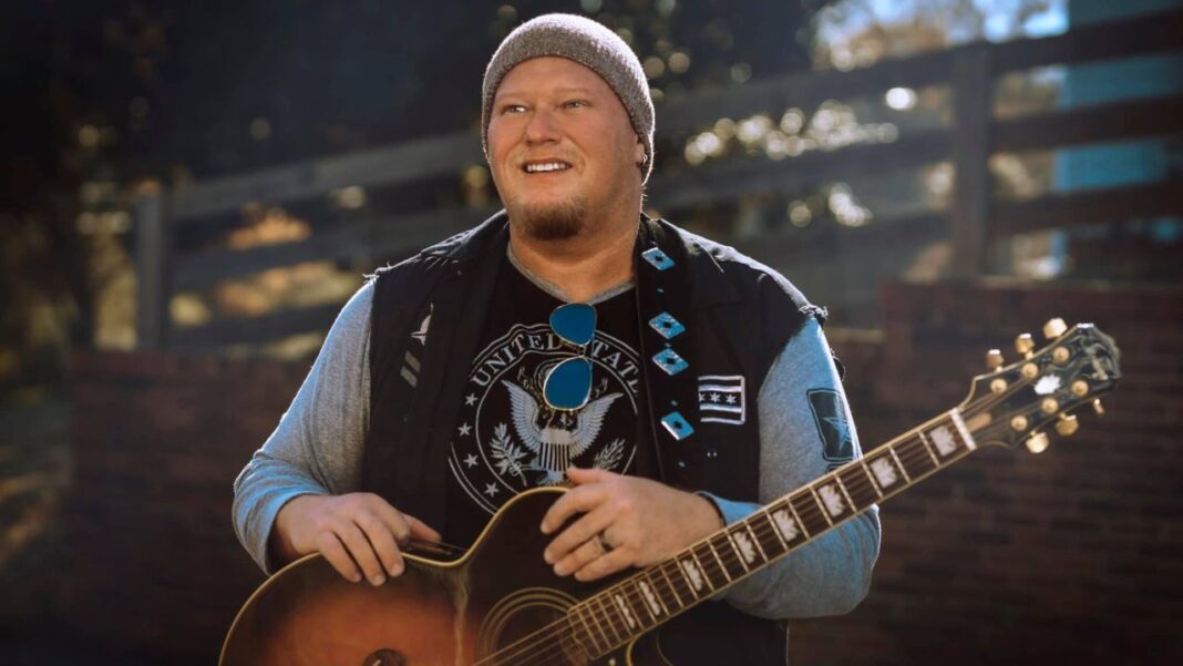 Hombre sonriente con gorro de punto, chaleco negro y camiseta gris sostiene una guitarra acústica frente a una valla de madera.