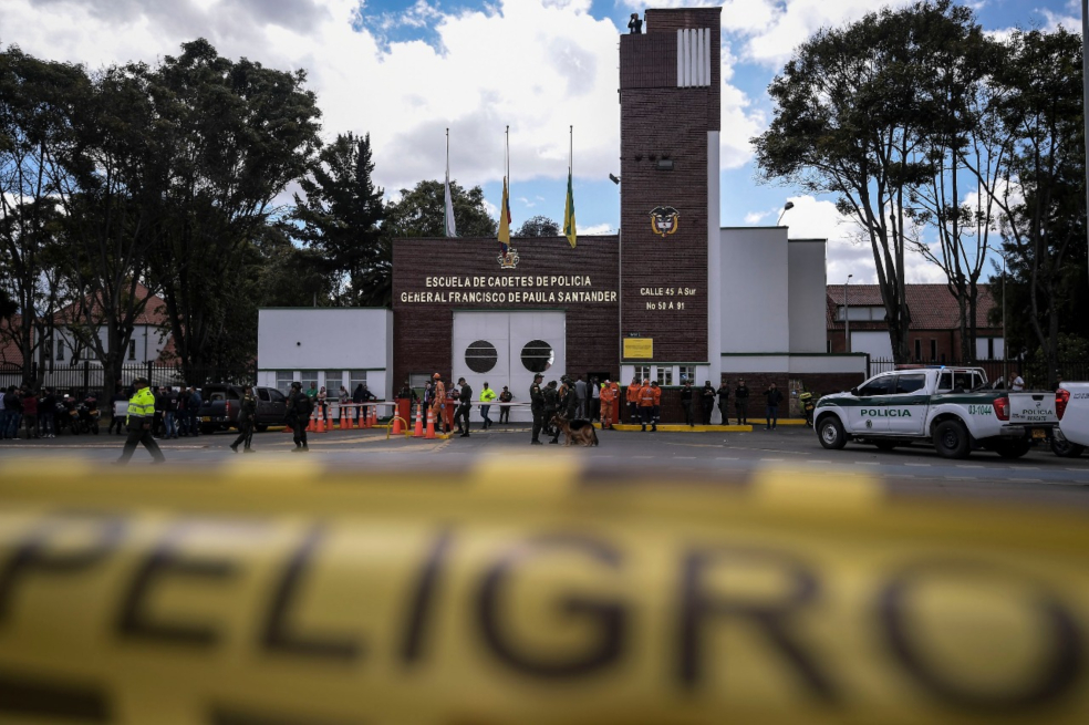 Fachada de la Escuela de Cadetes de Policía General Francisco de Paula Santander con personas reunidas, una camioneta policial y cinta amarilla de peligro en primer plano.