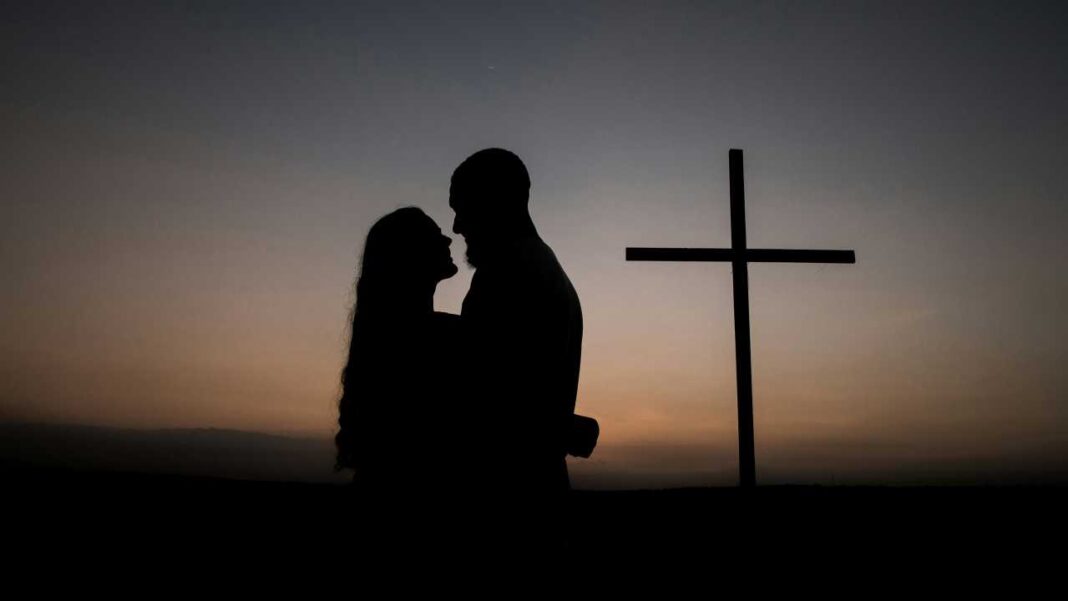 Silueta de una pareja de pie frente a frente junto a una gran cruz, bajo un cielo degradado al atardecer.