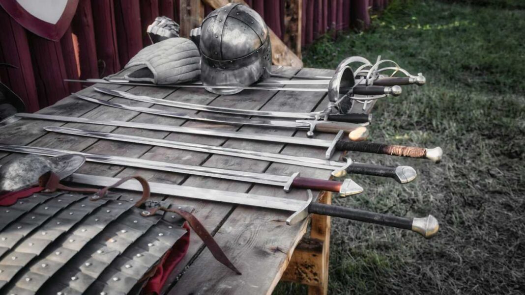 Varias espadas dispuestas sobre una mesa de madera junto a un casco metálico, una prenda acolchada y piezas de armadura, con césped y valla roja al fondo.
