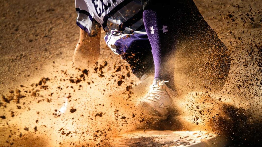 Jugador de béisbol deslizándose hacia una base mientras levanta una gran nube de tierra; se ven pierna, calcetín morado y zapato blanco.