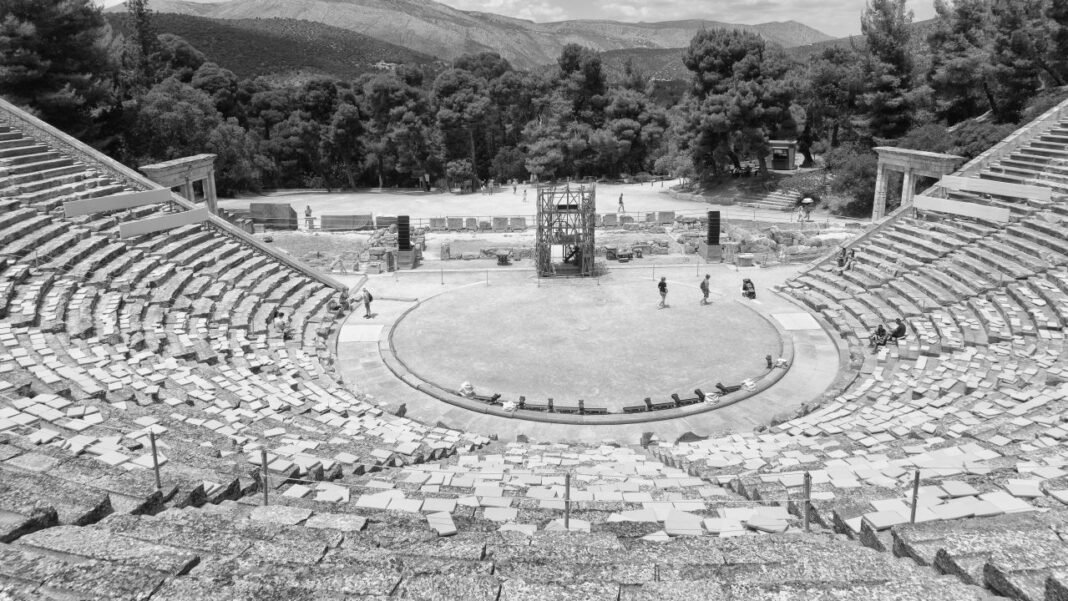 Vista en blanco y negro de un teatro antiguo al aire libre con gradas de piedra, orquesta circular, una estructura metálica central y árboles al fondo.