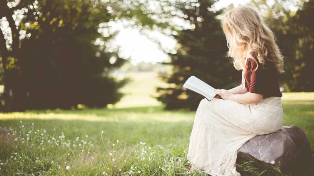 Joven rubia con cabello ondulado sentada sobre una roca en un campo verde, leyendo un libro abierto, con árboles desenfocados y flores blancas.
