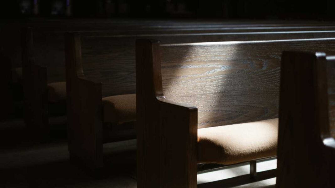 Filas de bancos de madera con asientos acolchados en una sala oscura; un haz de luz ilumina el respaldo de un banco.