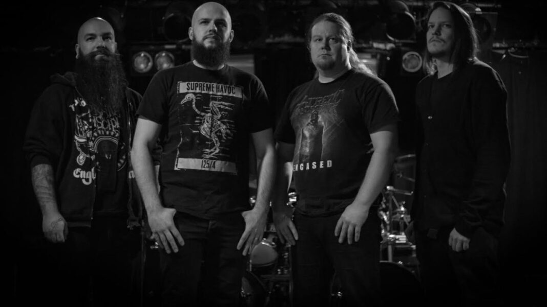 Cuatro hombres de pie en una habitación oscura en blanco y negro, con barbas y pelo largo, camisetas gráficas y una batería al fondo.