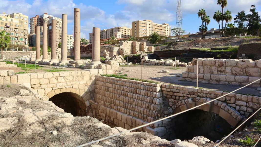 Ruinas de piedra con arcos y columnas antiguas frente a edificios modernos, palmeras y cielo azul con nubes.