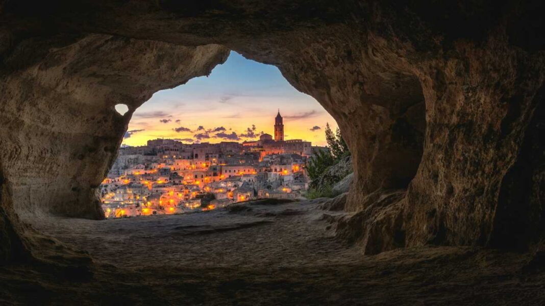 Vista de un pueblo iluminado al atardecer a través de una cueva de piedra, con torre, casas y cielo naranja y azul.