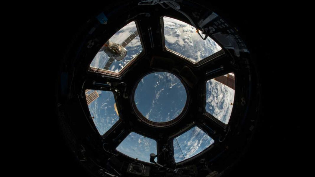 Vista desde una ventana circular de nave espacial con varios paneles, mostrando la Tierra, nubes y parte de un módulo exterior iluminado.
