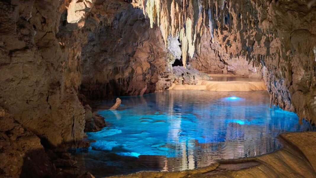 Cueva con formaciones rocosas colgantes y una laguna subterránea de agua turquesa iluminada, con reflejos y bordes de piedra.