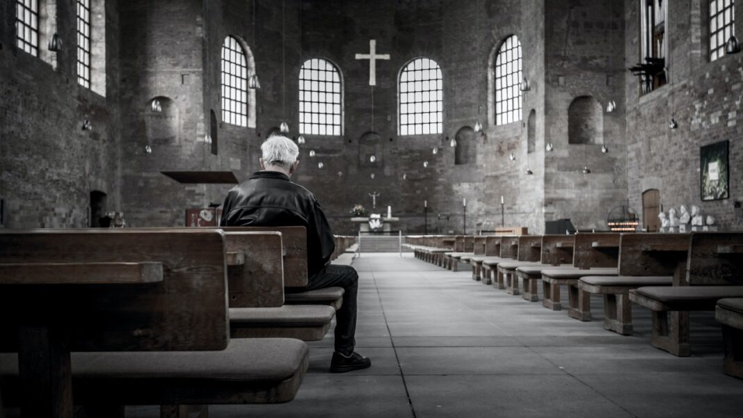 Persona sentada en un banco de madera dentro de una iglesia de piedra, mirando hacia el altar y una cruz.