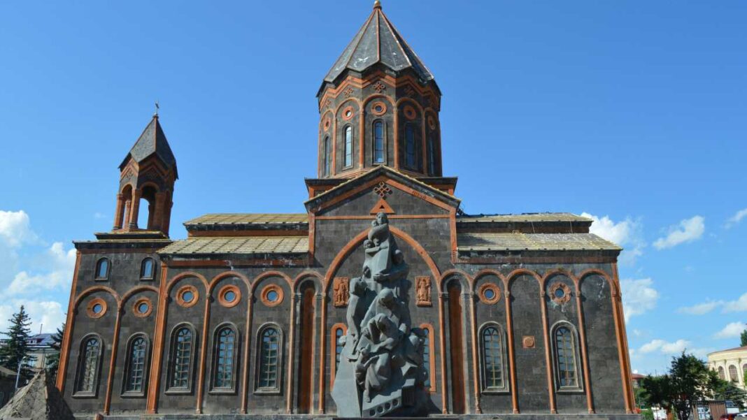 Gran iglesia de piedra oscura con cúpula cónica central, torre lateral, ventanas arqueadas y un monumento escultórico frente a la fachada bajo cielo azul.