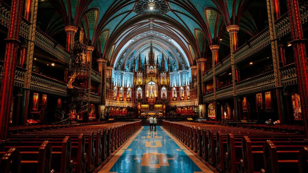 Interior de una iglesia con bóvedas azules estrelladas, columnas ornamentadas, bancos de madera y un pasillo central; dos personas de pie frente al altar.