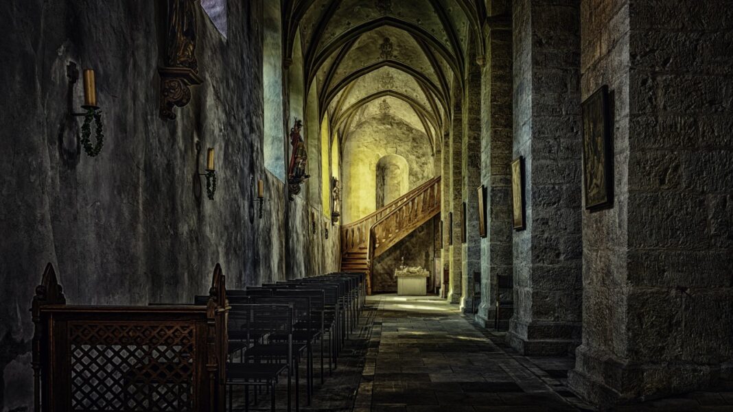 Interior de piedra con techo abovedado, sillas alineadas en un pasillo, candelabros en la pared y una escalera de madera al fondo.