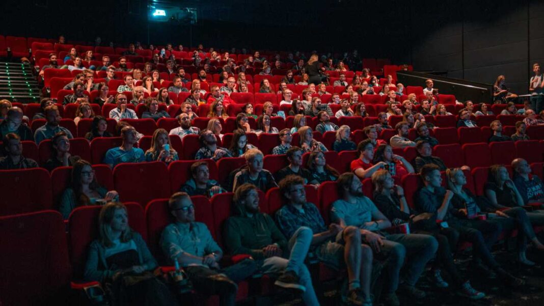 Personas de distintas edades sentadas en filas de butacas rojas en una sala oscura, mirando hacia el frente.
