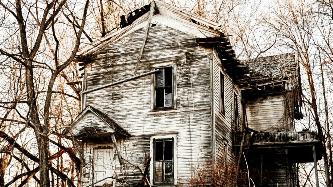 Casa de madera abandonada y deteriorada entre árboles sin hojas, con tejado dañado, ventanas oscuras y fachada desgastada.