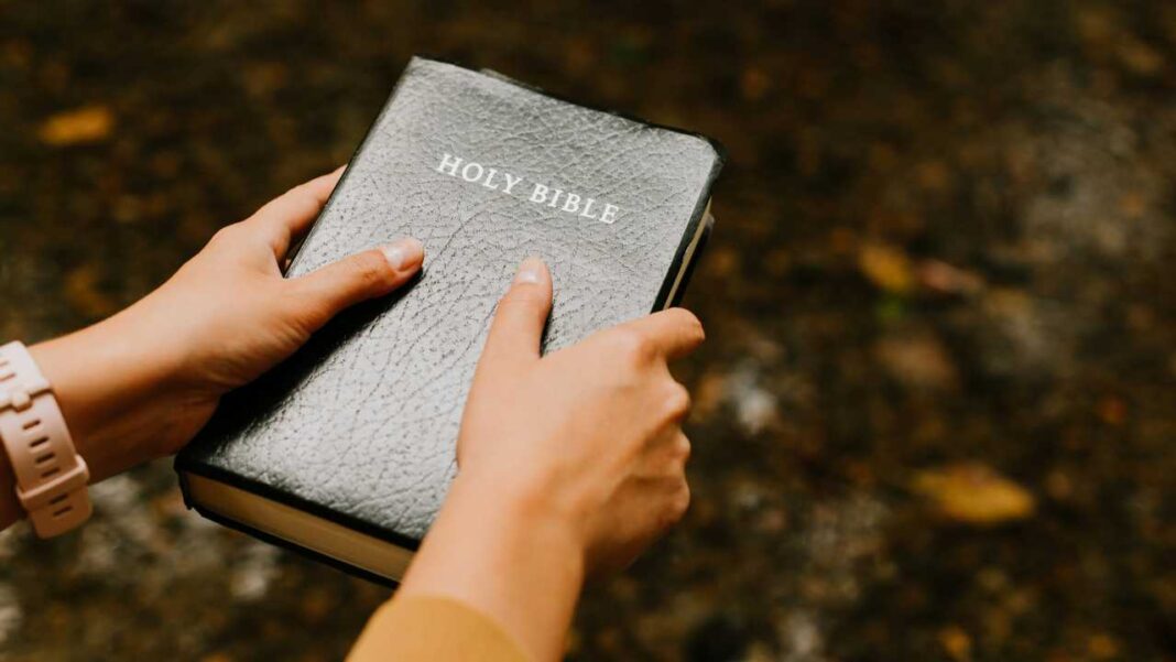 Dos manos sostienen una Biblia negra con la inscripción Holy Bible; una muñeca lleva un reloj rosa y el fondo está desenfocado.