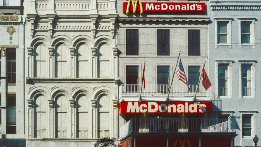 Edificio blanco ornamentado con ventanas arqueadas, letreros de McDonald's, banderas en un balcón y fachadas vecinas.