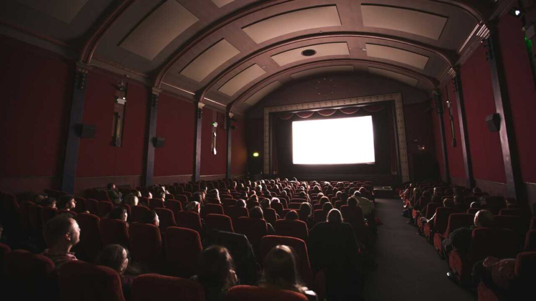 Sala de cine con paredes rojas, filas de butacas ocupadas por el público y una gran pantalla blanca luminosa al frente bajo un techo abovedado.