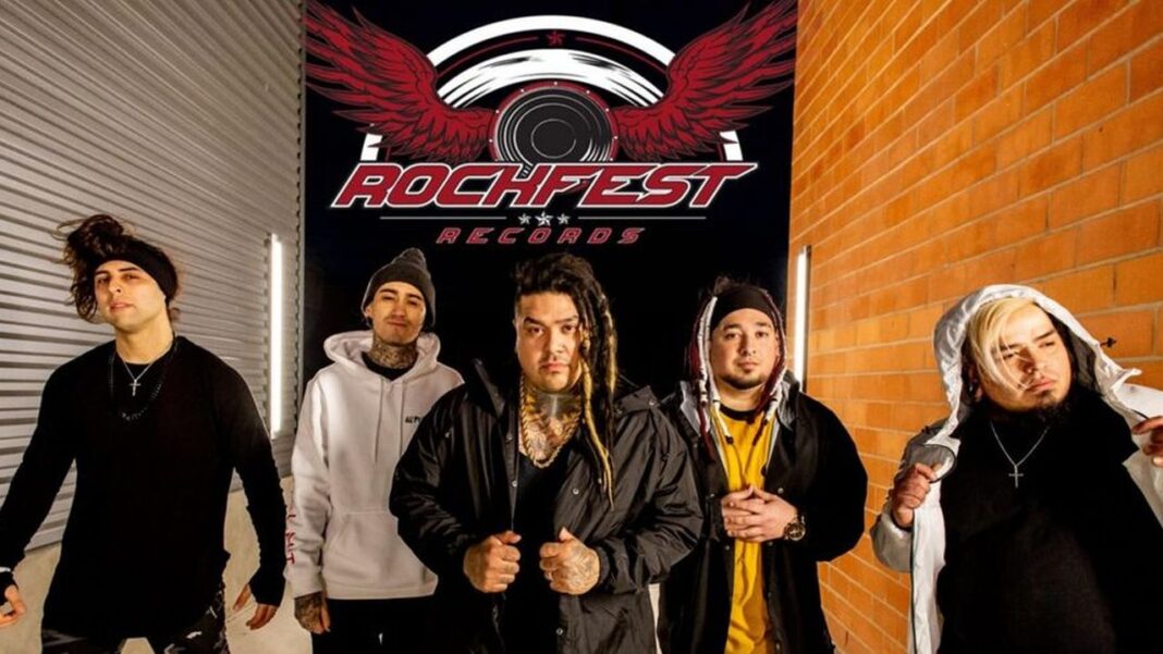 Cinco hombres de pie frente a un letrero negro con alas rojas y el texto Rochfest Records, junto a una persiana metálica y una pared de ladrillos.