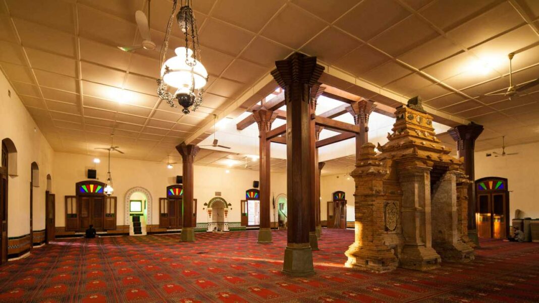 Interior de una sala amplia con columnas de madera, alfombra roja estampada, techo de panel, lámparas colgantes y una estructura de piedra ornamentada.