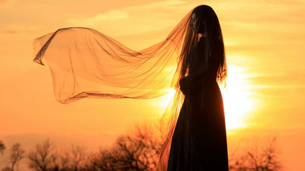 Silueta de una persona con vestido oscuro y velo transparente al viento frente a un cielo naranja de atardecer y árboles sin hojas.